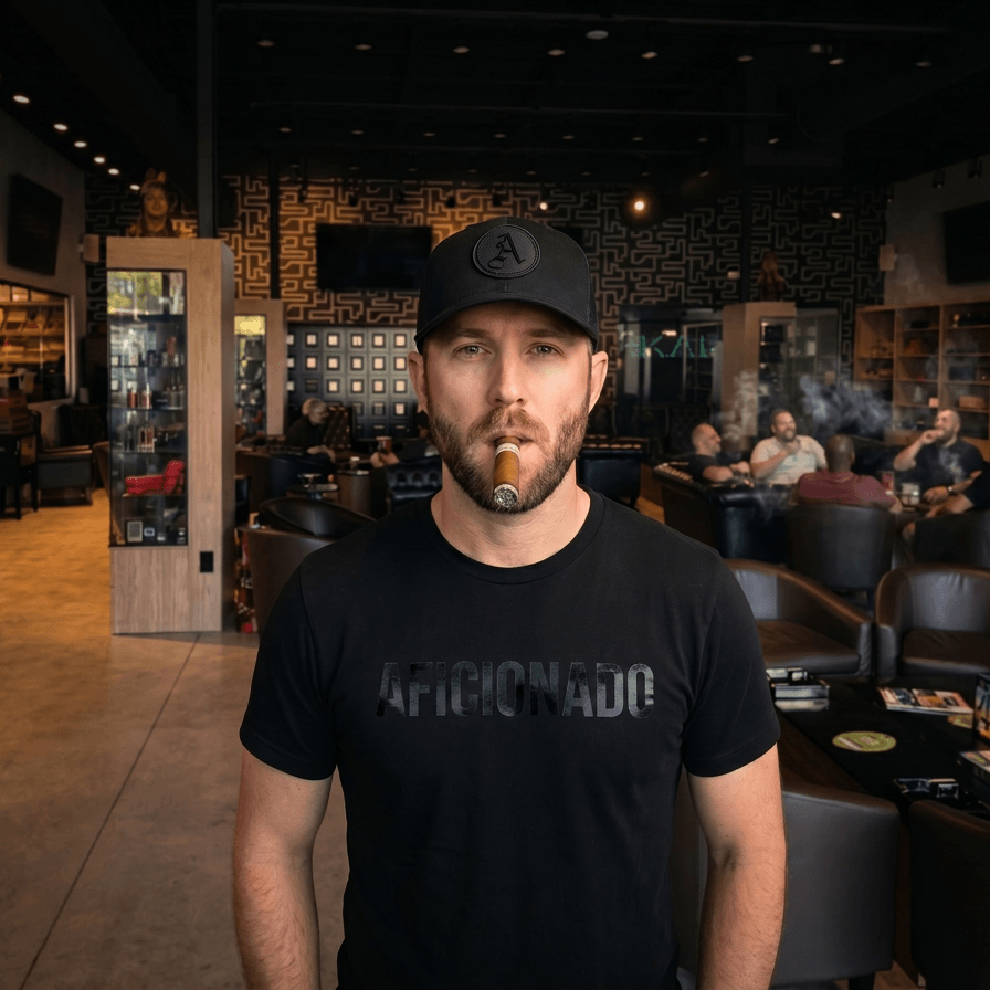 Black Curved Trucker with Black Aficionados Logo on White Male Wearing a Matching Tan Shirt Smoking a Cigar in Aficionados Lounge