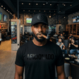 Black Curved Trucker with Black Aficionados Logo on Black Male Wearing a Matching Tan Shirt Smoking a Cigar in Aficionados Lounge